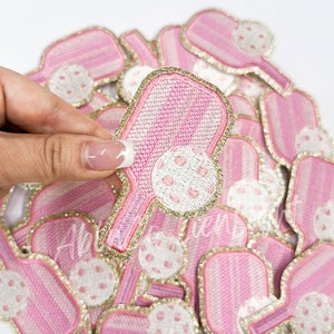 May include: A pile of pink and gold iron-on patches featuring a paddle and ball design. The patches have a striped pattern and white polka dots.