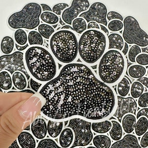 May include: A black and white paw print patch with sequins. The patch is surrounded by other paw print patches.