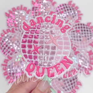 May include: A collection of pink sequined disco ball patches with the words "Dancing Queen" embroidered in a bright pink font. The patches feature a star design and are set against a white background.