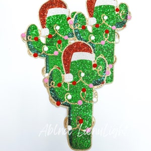 May include: Three green cactus ornaments with red Santa hats and white and pink Christmas lights. The ornaments are made of sequins and beads.
