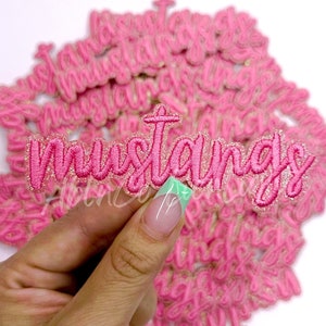 May include: Pink glitter iron-on patch with the word "Mustangs" in a cursive font.