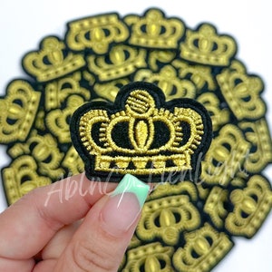 May include: A close-up of a gold embroidered crown patch with a black background. The patch is being held by a hand with light green nail polish.