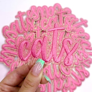 May include: A pile of pink and gold glitter iron-on patches with the word "cats" written in a cursive font.