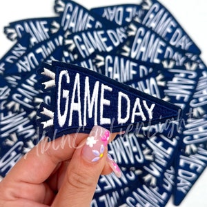 May include: A stack of navy blue felt pennants with white stitching that reads "GAME DAY".