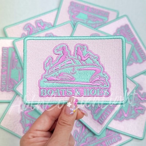 May include: A stack of pink and light blue embroidered patches with the text "Boats & Hoes" in white. The patches are square and have a white background with a pink outline.