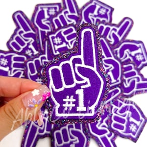May include: Purple foam finger with white glitter and the number "#1" in white. The finger is raised in the air and is surrounded by other foam fingers.