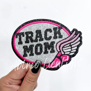 May include: Oval-shaped patch with the text "TRACK MOM" in black, glittery silver background, and a pink border. A pink and white wing design is on the right side. The patch is likely for apparel or accessories.