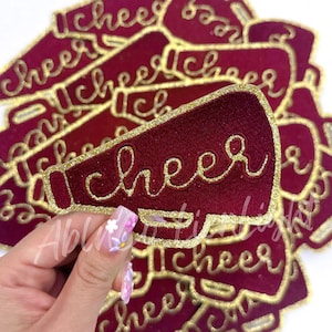 May include: A stack of maroon felt patches shaped like megaphones with gold glitter trim and the word "cheer" embroidered in gold glitter.
