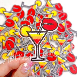 May include: A close-up of a red, yellow, and orange embroidered iron-on patch of a cocktail glass with a straw, a red lip, and a lemon slice. The patch is being held by a hand with colorful nail polish.