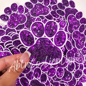 May include: A large group of purple paw print iron-on patches with white outlines. The patches are made of sequins and are perfect for adding a touch of sparkle to any project.