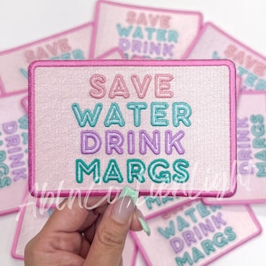 May include: Pink embroidered patch with the text "SAVE WATER DRINK MARGS" in pastel colors. The patch has a pink border and is rectangular. Several similar patches are visible in the background.