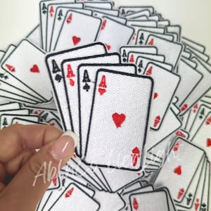 May include: A stack of white felt embroidered playing card patches with black outlines and red hearts. The patches are all aces.