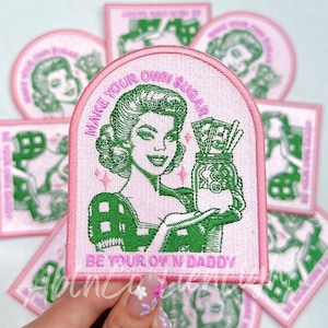May include: Pink and green embroidered iron-on patch with a retro woman holding a jar of money. The text reads "Make your own sugar, be your own daddy."