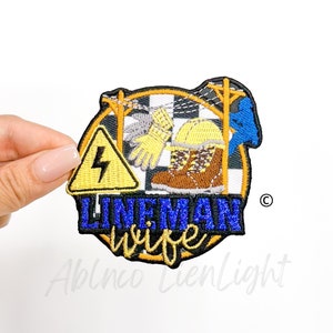 May include: Embroidered patch with the words "Lineman Wife" in white and blue script. The patch features a yellow hazard sign with a lightning bolt, work boots, a hard hat, and utility poles. The patch is surrounded by a gold border.