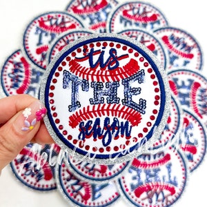 May include: A close-up of a white, round, baseball-themed patch with a silver glitter border. The patch features red and blue stitching and rhinestones. The text "'tis the season" is embroidered in red and blue thread.