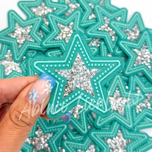 May include: A pile of teal blue fabric star-shaped patches with silver glitter in the center. The stars have a white outline and a dotted border.