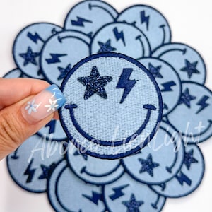 May include: A stack of blue denim iron-on patches with a smiling face, a lightning bolt, and a star. The star is glittery.