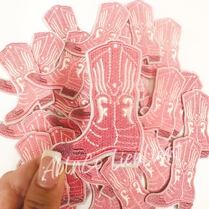 May include: A pile of pink embroidered iron-on patches shaped like cowboy boots. The boots have white stitching and a white star design.