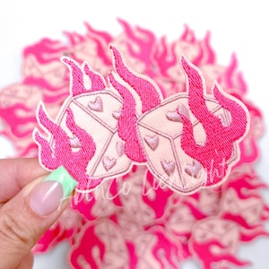 May include: Pink and white embroidered iron-on patch of two dice with hearts on each side, engulfed in pink flames.