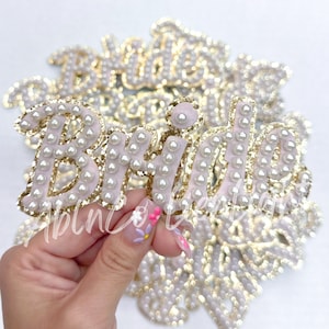 May include: A close-up of a pink fabric patch with the word "Bride" in gold glitter and white pearl embellishments. The patch is surrounded by other similar patches.