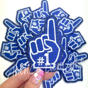 May include: A pile of blue and white foam finger patches with the number one and a hashtag. The patches are covered in glitter.