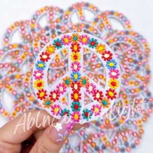 May include: A white felt peace sign with a colorful flower design. The peace sign is surrounded by other peace signs.