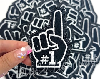 May include: A stack of black and white foam finger patches with the number one and the word "#1" embroidered in white. The patches are covered in glitter.