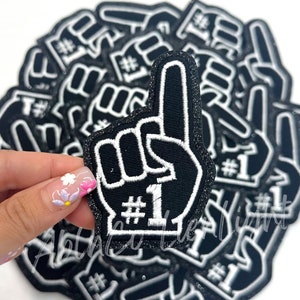 May include: A stack of black and white foam finger patches with the number one and the word "#1" embroidered in white. The patches are covered in glitter.