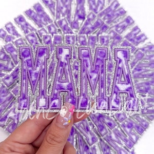 May include: Embroidered patch with the word "MAMA" in purple and white tie-dye style letters. The letters are outlined with silver glitter. A hand is holding the patch in front of a background of similar patches.