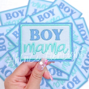 May include: Embroidered patch with the words "BOY MAMA" in light blue and teal. The patch is rectangular with a light blue background and a teal border. Other patches with the word "BOY" are in the background.