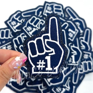 May include: Navy blue foam finger patches with a white outline and the number one. The patches have a glittery dark blue border. The patches are for sports fans and are a fun way to show support for a team.