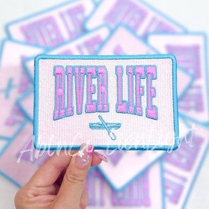May include: A white embroidered patch with a blue border. The patch says "River Life" in pink letters with a small blue and white rowing boat underneath.