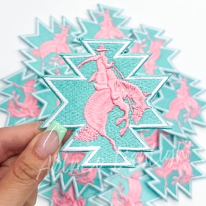 May include: A stack of turquoise blue and pink embroidered iron-on patches featuring a cowboy riding a bucking horse. The patches are in the shape of a starburst.