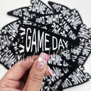 May include: A close-up of a black felt patch with white embroidered text that reads "GAME DAY". The patch is shaped like a pennant and is held in a hand with painted nails.