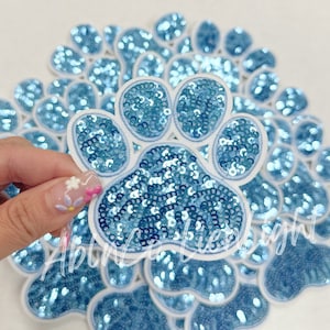 May include: A pack of 20 light blue paw print iron-on patches. Each patch is made of sequins and has a white outline.