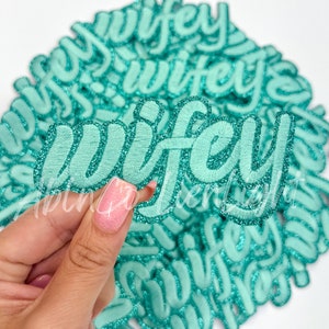 May include: A pile of teal blue iron-on patches with the word "wifey" in a glittery font.