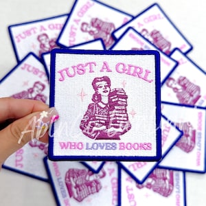 May include: A white embroidered patch with a blue border. The patch features a woman holding a stack of books and the text "Just a girl who loves books".