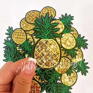 May include: A pile of gold and green pineapple-shaped iron-on patches. The pineapples are made of sequins and have green leaves.