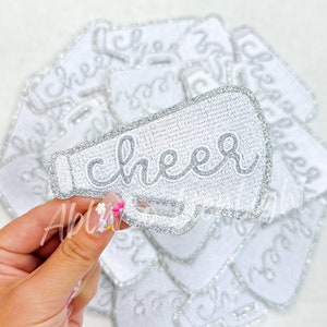 May include: A white iron-on patch with silver glitter trim shaped like a megaphone with the word "cheer" written in a silver cursive font.