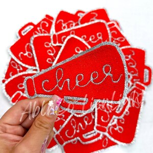 May include: A stack of red felt patches shaped like a megaphone with a silver glitter outline and the word "cheer" written in silver glitter.