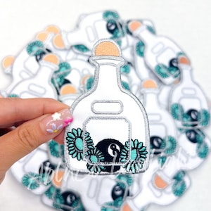 May include: A white bottle with a silver lid and a black 8-ball inside. The bottle is surrounded by turquoise flowers. The bottle is a patch.