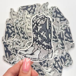 May include: Embroidered cowboy boot patch in shades of gray and white, adorned with silver-colored rhinestones. The boot features intricate floral patterns and is surrounded by similar patches.
