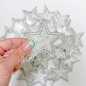 May include: A pile of silver glitter star-shaped iron-on patches. The patches are approximately 2 inches in diameter.