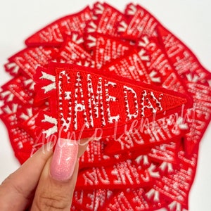 May include: A stack of red felt pennant patches with white embroidered text that reads "Game Day" with rhinestones. The patches are approximately 3 inches long and 2 inches wide.
