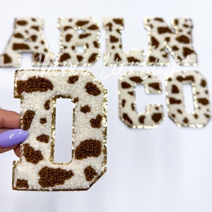 May include: Cow print felt letters in brown and white with gold trim. The letters spell out "ABLNCO".