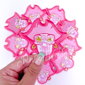 May include: Pink embroidered patch featuring a skull wearing a pirate hat. The skull has star-shaped eyes and gold glitter accents. The patch is held by a hand with a light green manicure. Other patches are in the background.