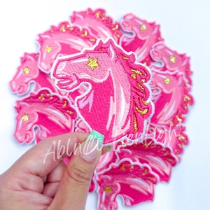 May include: A collection of pink horse head patches with gold accents. The patches feature a star on the forehead and are embroidered with a white outline. The horse's mane and details are highlighted with gold thread.