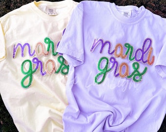 May include: Two t-shirts with the words "Mardi Gras" in green, purple, and gold sequins. The shirts are a light yellow and a light purple.
