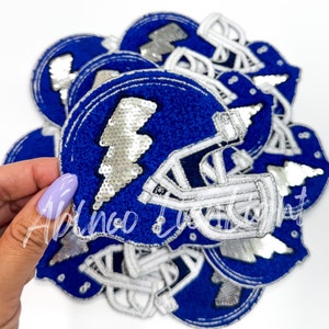 May include: A stack of blue and white felt iron-on patches featuring a football helmet with a silver lightning bolt made of sequins.