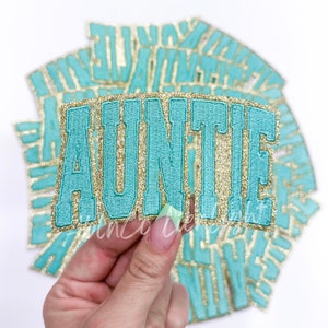 May include: A teal blue and gold glitter iron-on patch that says "AUNTIE".